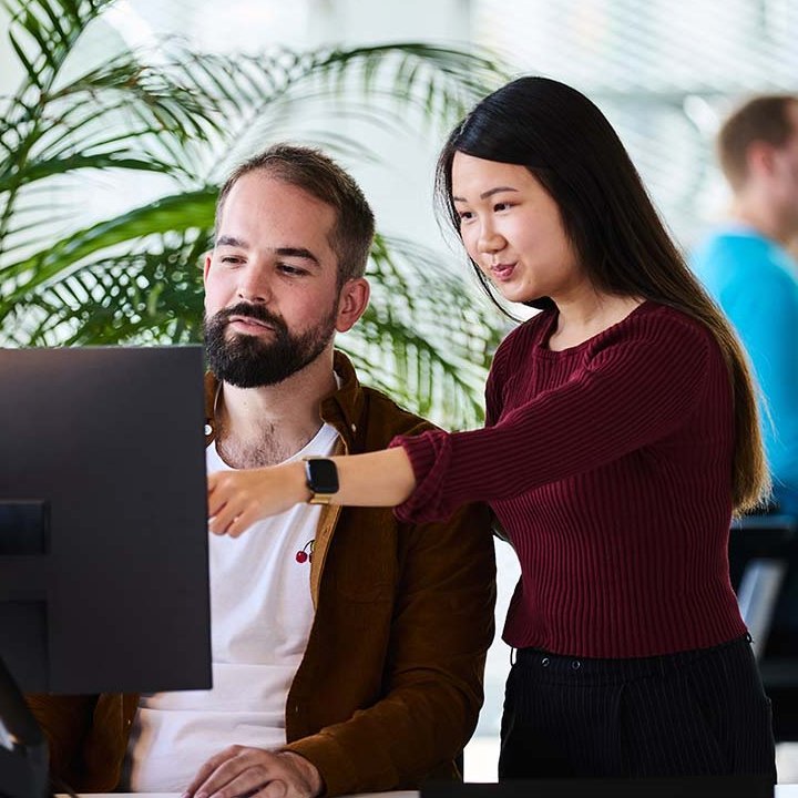 Eine lise-Entwicklerin zeigt einem anderen etwas auf seinem Computer und im Hintergrund unterhalten sich weitere Mitarbeiter