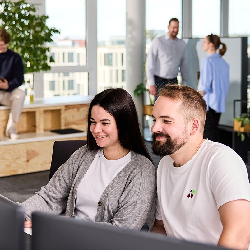 Zwei lise-Entwickler*innen betrachten gemeinsam ihr Ergebnis auf einem Computer und im Hintergrund sind weitere Mitarbeiter