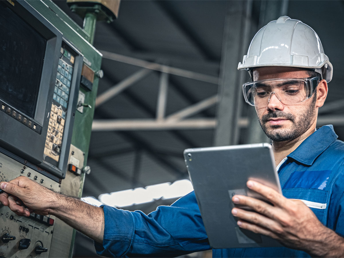 Ein Mann mit Schutzhelm drückt einen Knopf an einer Industriemaschine und hält ein Tablet in der Hand.