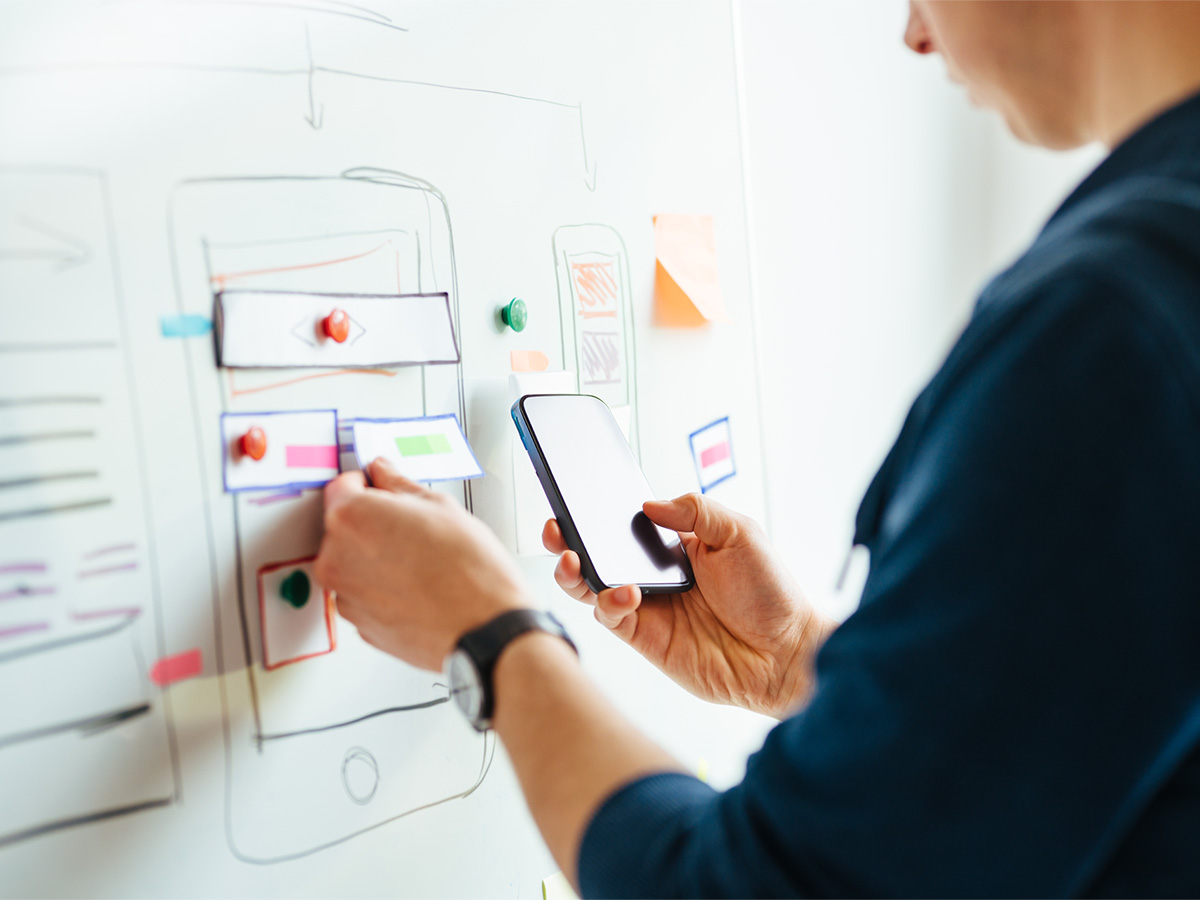 Eine Frau pinnt an eine Whiteboard Zettel und hält ein Handy in der Hand. 