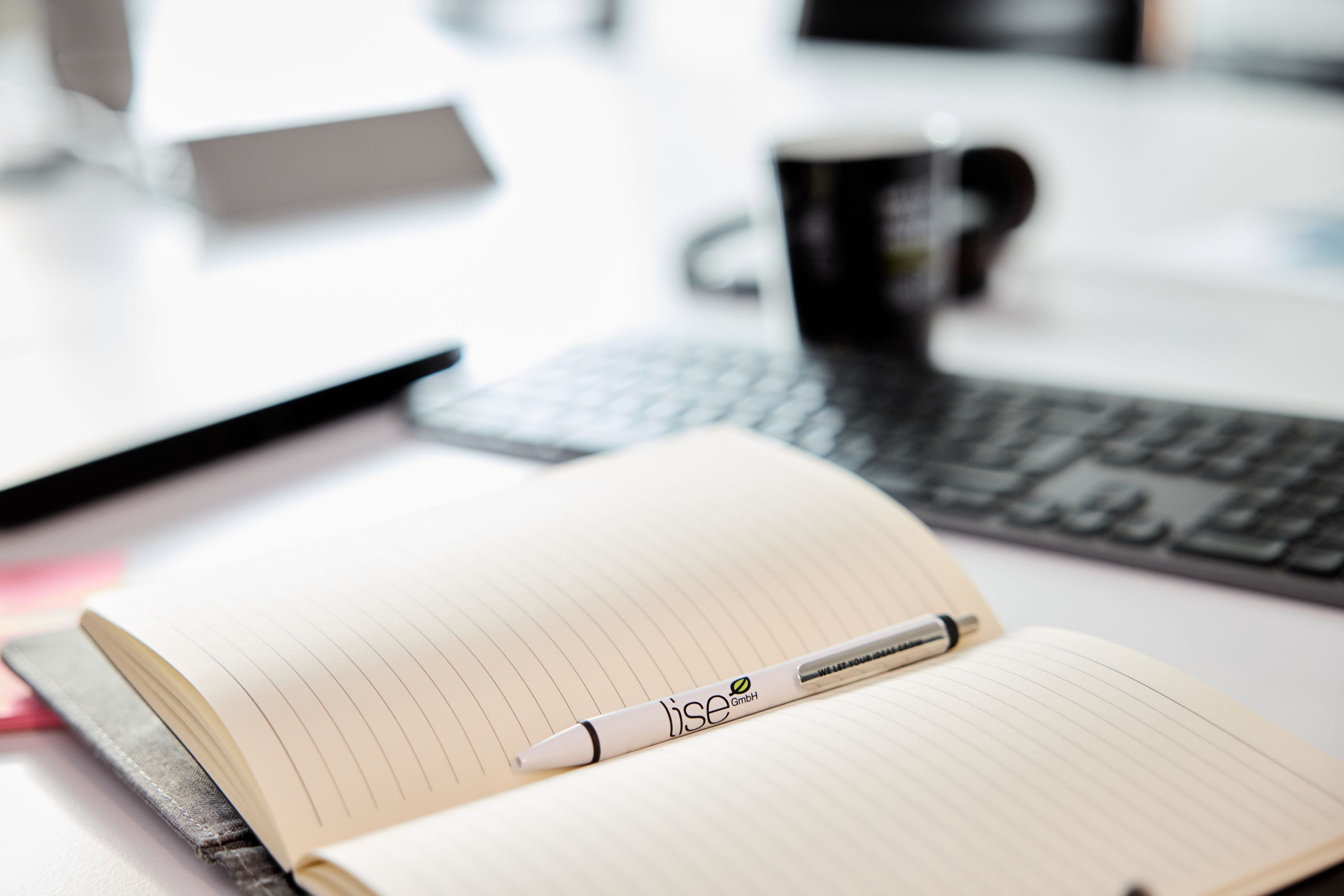 Ein aufgeschlagenes leeres Notizbuch liegt auf dem Tisch mit einem Stift, einer Tastatur und einer Tasse im lise-Design