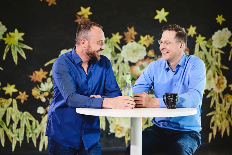 Die zwei Geschäftsführer der lise sitzen an einem erhöhten Tisch und unterhalten sich lachend vor einem bunten Hintergrund